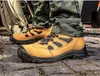 Close-up of work boots on a cobblestone surface, with camouflage pants and outdoor gear in the background. Voir les images du produit
