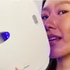 A woman is holding a white LED face mask to her face, with the blue light from the mask visible. Anteprima dell’immagine prodotto