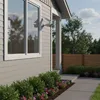 A security camera is mounted on the side of a house next to a window, with a walkway and garden in the foreground. Produktbild-Vorschau