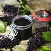 A jar of dark liquid, likely elderberry syrup, surrounded by fresh elderberries and leaves on a wooden surface. Produktbild-Vorschau