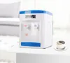 A white and blue tabletop water dispenser is shown on a white table, with a blurred background of a living room. Náhled obrázku produktu