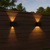 Two black cube-shaped outdoor wall lights mounted on a dark wooden fence, casting warm yellow light upwards and downwards. Náhľad obrázku produktu