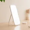 A white LED makeup mirror with a stand is placed on a wooden surface, with a blurred background of a wall and some cosmetic items. Podgląd zdjęcia produktu