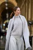 A woman wearing a light gray knitted sweater and skirt, with a matching knitted shawl draped over her shoulders, walking outdoors in an arched walkway. Produktbild-Vorschau