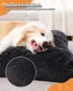 A dog is lying on a dark gray plush dog bed. The background shows a couch and a blanket. Produktbild-Vorschau