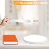 A hand holds a thin, flat, circular object, demonstrating its slim profile. Next to it on a table are an orange book and a white circular light fixture. Voir les images du produit