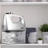 A silver electric hand mixer with its accessories stored in a clear container sits on a shelf next to stacked cups and a potted plant. Produktbild-Vorschau
