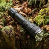 A black flashlight rests on a mossy, wooded surface with fallen leaves. Podgląd zdjęcia produktu