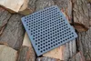 A square cast iron grate with many holes rests on a pile of chopped wood. Produktbild-Vorschau