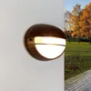 A wall-mounted outdoor light fixture is shown on a white wall, with a blurred background of trees in autumn colors and a paved walkway. Podgląd zdjęcia produktu