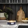 A lit Woodwick candle sits on a marble cutting board on a kitchen counter, with a plant and cutting boards in the background. Podgląd zdjęcia produktu