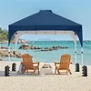 A navy blue pop-up canopy tent set up on a sandy beach with two wooden chairs and a small table. Produktbild-Vorschau