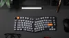 A top-down view of a split ergonomic keyboard on a black desk, with a mouse, glasses, and a pen visible. Produktbild-Vorschau