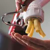 A hand is holding a pasta maker attachment on a red stand mixer, with fresh pasta being extruded into the hand. Náhled obrázku produktu