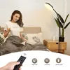 A woman is sitting in bed reading a book and drinking from a mug, with a floor lamp and a nightstand next to the bed. The bottom of the image shows a remote control and icons representing different modes. Produktbild-Vorschau