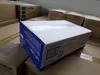 Several cardboard boxes are stacked in a warehouse or storage area. The focus is on a blue and white Brother branded box in the foreground. Voir les images du produit