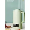 A light green soy milk maker is displayed on a white table with some food items in the foreground. The background is a light green wall with vertical paneling. Anteprima dell’immagine prodotto