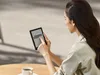 A woman is sitting at a table outdoors, reading an e-reader. Produktbild-Vorschau