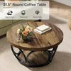 A round coffee table with a wood-grain top and a black metal base, decorated with a vase of flowers and two books. Anteprima dell’immagine prodotto