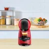 A red and black Krups Dolce Gusto Piccolo XS coffee machine is shown on a wooden countertop in a kitchen setting. Behind the machine, shelves hold jars of food items and a bowl of fruit. Anteprima dell’immagine prodotto