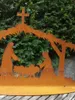 A metal nativity scene with a cross on top, set against a backdrop of green foliage and a stone wall. Anteprima dell’immagine prodotto