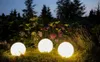 Three illuminated spherical garden lights are placed on the ground in a garden at dusk, surrounded by plants and trees. Anteprima dell’immagine prodotto