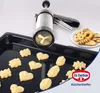 A cookie press and freshly made cookies on a baking sheet, with a plate of finished cookies in the background. Anteprima dell’immagine prodotto