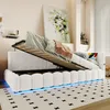 A white upholstered bed frame with the mattress lifted, revealing storage space underneath. The bed has LED lighting around the base. A painting hangs on the wall behind it. Voir les images du produit