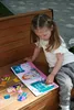 A young girl is playing with a magnetic doll dress-up game on a wooden bench. Produktbild-Vorschau