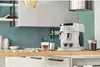 A white De Longhi coffee machine is on a kitchen counter, with two cups of coffee and a jar of coffee beans. Produktbild-Vorschau