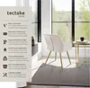 A beige upholstered chair with gold legs is placed in a room with a large window overlooking a forest. A small side table with a mug and book is next to the chair. Produktbild-Vorschau