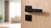 A black door handle set is mounted on a light wood door. The background is blurred, showing a room with furniture and curtains. Produktbild-Vorschau