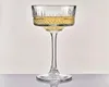 A single champagne coupe glass filled with a pale yellow liquid, set against a plain white background with a reflective surface below. Náhľad obrázku produktu