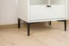 Close-up of the bottom of a white cabinet with black metal legs, resting on a light wood floor. Produktbild-Vorschau