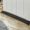 A dark gray fabric draft stopper lies on a wooden floor in front of a white door. Produktbild-Vorschau