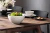 A white textured serving bowl filled with salad sits on a wooden dining table, next to a white bowl and a small plate with food. A vase of white flowers is visible in the background. Podgląd zdjęcia produktu