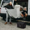 A couple is setting up camp next to a van, with a cooler on the ground. Produktbild-Vorschau
