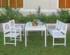 A white wooden dining set with a table and chairs is placed on a grassy lawn in front of a building with green bushes and yellow flowers. Produktbild-Vorschau