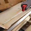 A gloved hand is using a red tape measure to measure a wooden plank. Produktbild-Vorschau