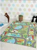 A children's play rug with a road and town design is laid out on a floor, with toys and stuffed animals around it. Produktbild-Vorschau