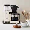 A coffee maker with a carafe of coffee, a croissant, and a cup of coffee on a table. Produktbild-Vorschau