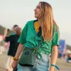 A woman at an outdoor event, possibly a festival, wearing a green shirt and holding a small green bag. Produktbild-Vorschau