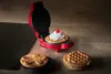 A red mini waffle maker is open and topped with whipped cream and a cherry, sitting on a wooden counter next to a black induction hob. Two other waffles are visible on the counter in the foreground. Produktbild-Vorschau