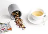 A grey canister of loose tea spilled onto a white surface, next to a white teacup filled with light yellow tea. Náhled obrázku produktu