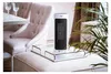 A white and black tower heater sits on a mirrored tray on a plush ottoman, next to a tufted chair and a dining table with wine glasses. Podgląd zdjęcia produktu