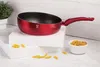 A red wok pan is displayed on white blocks, with a small bowl of pasta and some scattered pasta in the foreground. Produktbild-Vorschau