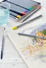 A watercolor pencil set, a glass of water, and a paintbrush are arranged on a wooden surface next to a sketchpad with a drawing. Produktbild-Vorschau