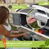 A woman is demonstrating the features of a two-story rabbit hutch, pulling out a drawer. The hutch is set up outdoors in a grassy area with a stone wall in the background. There are also some fruits visible in one of the drawers. Produktbild-Vorschau