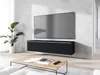 A modern living room with a TV mounted on the wall above a black media console. A coffee table with a book and remote is in front of the console, and a potted plant is to the left. Náhľad obrázku produktu