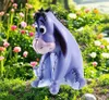 A purple Eeyore figurine sits in a garden surrounded by flowers and greenery. Produktbild-Vorschau
